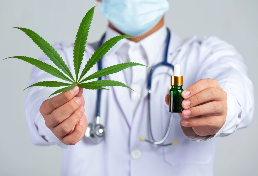A doctor wearing a lab coat, stethoscope, and face mask holds a cannabis leaf in one hand and a small THC dropper bottle in the other, both extended toward the camera.